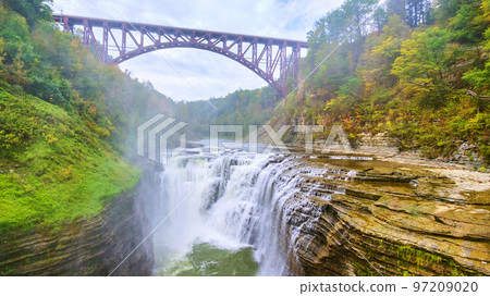 Waterfall cutting through cliffs from drone with steel maroon bridge overhead 97209020
