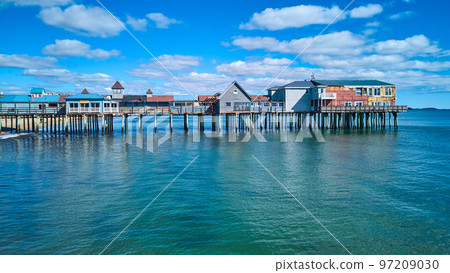 Aerial side profile of long old wood pier covered in shops in Maine with ocean and blue sky Aerial side profile of long old wood pier covered in shops in Maine with ocean and blue sky 97209030