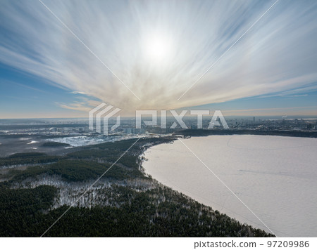 Snow-covered forest on lake shore with ice at sunset and the city on horizon, auerial view Snow-covered forest on lake shore with ice at sunset and the city on horizon, auerial view 97209986