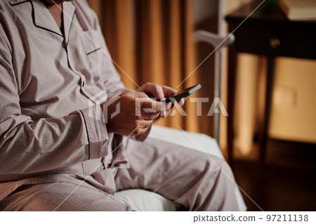 Cropped image of elderly man in pajamas texting friends when sitting on bed before going to sleep Cropped image of elderly man in pajamas texting friends when sitting on bed before going to sleep 97211138