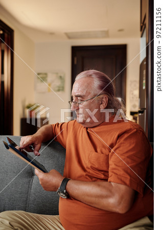Portrait of senior man in glasses reading article on tablet computer when spending evening at home 97211156