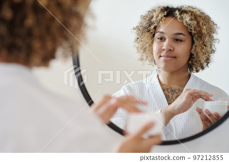 Young gorgeous woman looking in mirror while going to apply cream on her face Young gorgeous woman looking in mirror while going to apply cream on her face 97212855