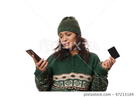 Charming worried woman in green sweater and hat, makes online bank payment, using a mobile app on her cell phone, worrying about possible failure of transaction due to lack of cash on credit card 97213184