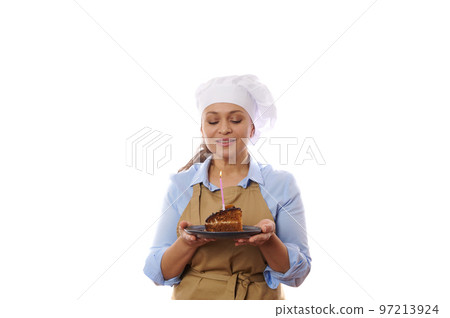 Happy woman pastry chef holding a plate with chocolate cake, making cherished wish and blowing out candle, celebrates birthday or anniversary, over white background. Confectionery and baking concept Happy woman pastry chef holding a plate with chocolate cake, making cherished wish and blowing out candle, celebrates birthday or anniversary, over white background. Confectionery and baking concept 97213924