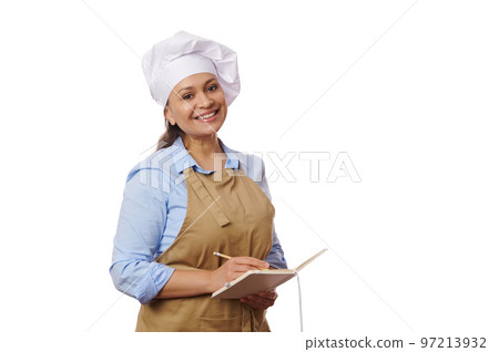Latin American woman, baker confectioner in white chef's hat and beige apron, writing on notepad, smiling a beautiful toothy smile looking at camera. Baking Culinary. Work in food and drink industry Latin American woman, baker confectioner in white chef's hat and beige apron, writing on notepad, smiling a beautiful toothy smile looking at camera. Baking Culinary. Work in food and drink industry 97213932