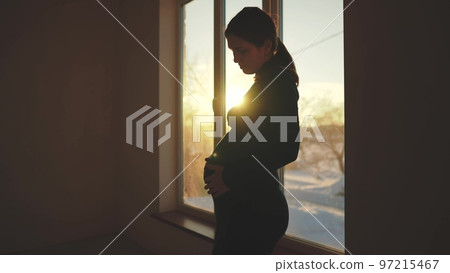 woman pregnant. motherhood a pregnancy concept. 40 year old pregnant woman stands in a dress by sunlight the window holding her stomach silhouette. silhouette of a girl in by the window 97215467