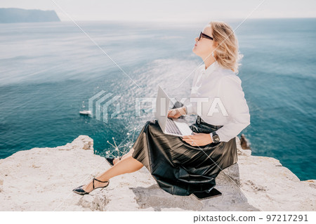 Digital nomad, Business woman working on laptop by the sea. Pretty lady typing on computer by the sea at sunset, makes a business transaction online from a distance. Freelance, remote work on vacation 97217291