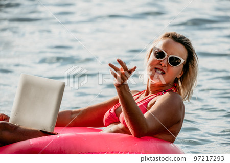 Woman works on laptop in sea. Freelancer, young blond woman in sunglases floating on an inflatable big pink donut with a laptop in the sea at sunset. Freelance, travel and holidays concept. 97217293