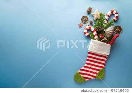 Christmas greeting with christmas gift sock, fir tree branches, Christmas candy cane, baubles, candy canes on blue background. Flat lay. Copy space 97219403