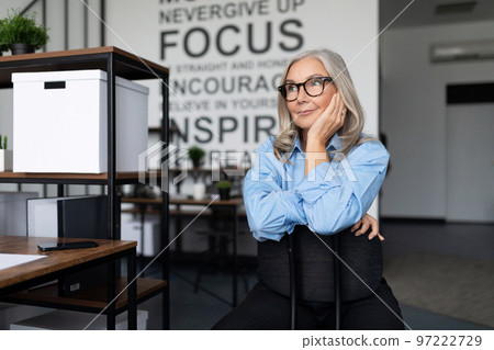 caucasian senior businesswoman in a blue shirt and glasses sits thinking in the office caucasian senior businesswoman in a blue shirt and glasses sits thinking in the office 97222729