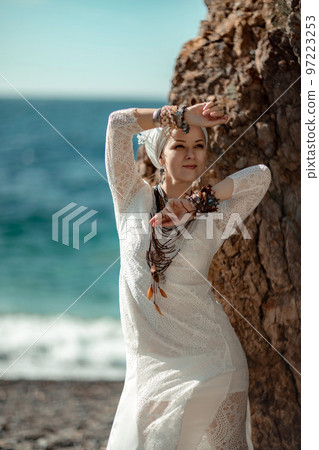 Middle aged woman looks good with blond hair, boho style in white long dress on the beach decorations on her neck and arms. Middle aged woman looks good with blond hair, boho style in white long dress on the beach decorations on her neck and arms. 97223253