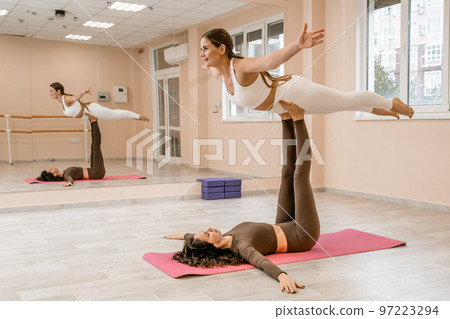 Two women do acroyoga on a light floor near a large mirror in a yoga studio. They are wearing leggings and tops. 97223294