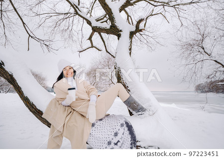A beautiful girl with a beige cardigan and a white hat enjoying drinking tea in a snowy winter forest near a lake 97223451