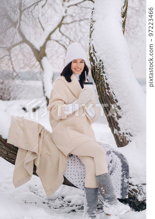 A beautiful girl with a beige cardigan and a white hat enjoying drinking tea in a snowy winter forest near a lake 97223466