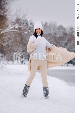 A beautiful girl in a beige cardigan and a white hat with a glass of tea enjoys a snowy embankment by the lake A beautiful girl in a beige cardigan and a white hat with a glass of tea enjoys a snowy embankment by the lake 97223705