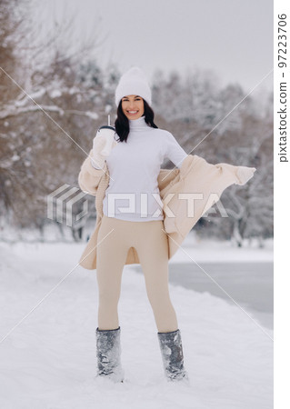 A beautiful girl in a beige cardigan and a white hat with a glass of tea enjoys a snowy embankment by the lake A beautiful girl in a beige cardigan and a white hat with a glass of tea enjoys a snowy embankment by the lake 97223706