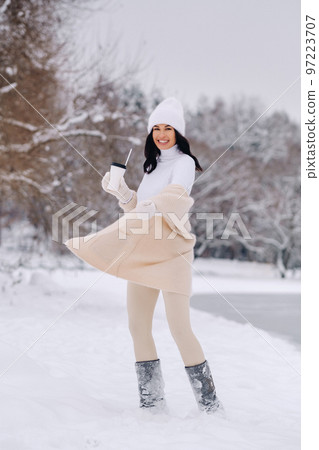 A beautiful girl in a beige cardigan and a white hat with a glass of tea enjoys a snowy embankment by the lake 97223707