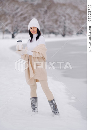 A beautiful girl in a beige cardigan and a white hat with a glass of tea enjoys a snowy embankment by the lake 97223742