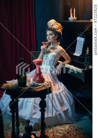 Cocktail evening. Portrait of young beautiful girl in image of medieval person in elegant white dress sitting at the piano. Comparison of eras, beauty, history, art Cocktail evening. Portrait of young beautiful girl in image of medieval person in elegant white dress sitting at the piano. Comparison of eras, beauty, history, art 97223828