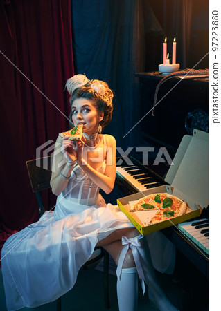 Portrait of young beautiful girl in image of medieval person in elegant white dress sitting at the piano and eating pizza. Comparison of eras, beauty, history, art Portrait of young beautiful girl in image of medieval person in elegant white dress sitting at the piano and eating pizza. Comparison of eras, beauty, history, art 97223880