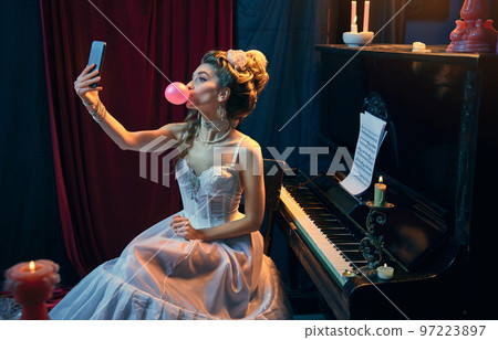 Bubblegum. Portrait of young beautiful girl in image of medieval person in elegant white dress sitting at the piano and taking selfie with phone. Comparison of eras, beauty, history, art Bubblegum. Portrait of young beautiful girl in image of medieval person in elegant white dress sitting at the piano and taking selfie with phone. Comparison of eras, beauty, history, art 97223897