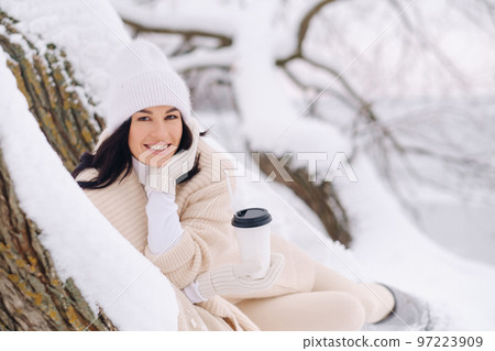 A beautiful girl with a beige cardigan and a white hat enjoying drinking tea in a snowy winter forest near a lake 97223909
