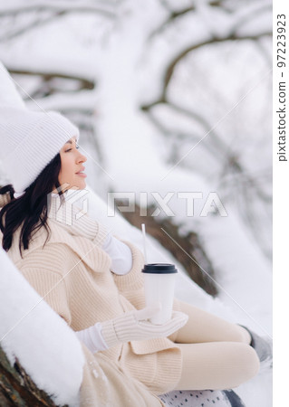 A beautiful girl with a beige cardigan and a white hat enjoying drinking tea in a snowy winter forest near a lake 97223923