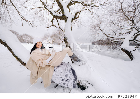 A beautiful girl with a beige cardigan and a white hat enjoying drinking tea in a snowy winter forest near a lake A beautiful girl with a beige cardigan and a white hat enjoying drinking tea in a snowy winter forest near a lake 97223947
