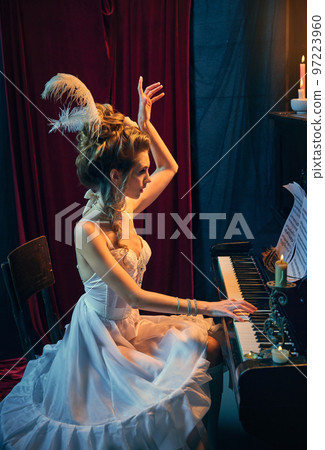 Elegance and tenderness. Portrait of beautiful young woman in image of medieval person in white dress sitting at piano and playing. Comparison of eras, beauty, history, art 97223960