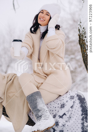 A beautiful girl with a beige cardigan and a white hat enjoying drinking tea in a snowy winter forest near a lake A beautiful girl with a beige cardigan and a white hat enjoying drinking tea in a snowy winter forest near a lake 97223966
