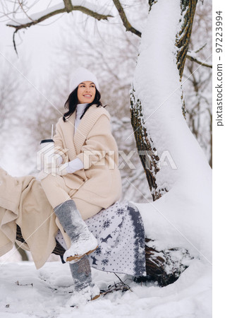 A beautiful girl with a beige cardigan and a white hat enjoying drinking tea in a snowy winter forest near a lake A beautiful girl with a beige cardigan and a white hat enjoying drinking tea in a snowy winter forest near a lake 97223994