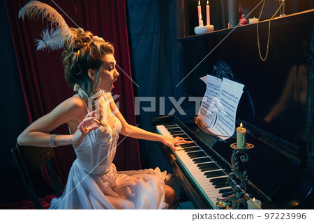 Coquettish look. Portrait of young beautiful woman in image of medieval person sitting at the piano in elegant white dress and playing. Comparison of eras, beauty, history, art 97223996