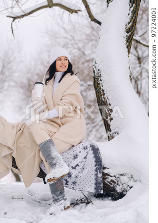 A beautiful girl with a beige cardigan and a white hat enjoying drinking tea in a snowy winter forest near a lake A beautiful girl with a beige cardigan and a white hat enjoying drinking tea in a snowy winter forest near a lake 97224001
