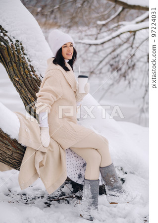 A beautiful girl with a beige cardigan and a white hat enjoying drinking tea in a snowy winter forest near a lake 97224031