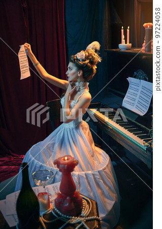 Young tender girl in image of medieval person in elegant white dress sitting at the piano and learning new music composition. Comparison of eras, beauty, history, art 97224058