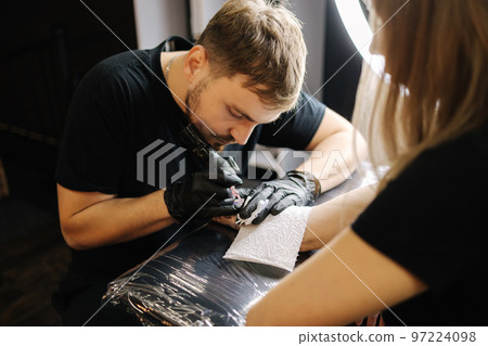 Tattoo master is tattooing a woman's hand. Wireless tattoo machine, safety and hygiene at work. Close-up of tattoo artist work. Tattoo salon Tattoo master is tattooing a woman's hand. Wireless tattoo machine, safety and hygiene at work. Close-up of tattoo artist work. Tattoo salon 97224098