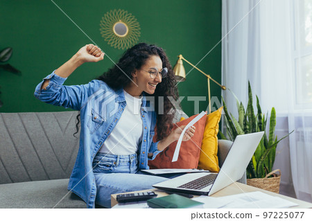 Happy hispanic woman at home doing paperwork and calculating household budget, sitting on couch in living room and using laptop at work, holding hand up victory and triumph gesture. Happy hispanic woman at home doing paperwork and calculating household budget, sitting on couch in living room and using laptop at work, holding hand up victory and triumph gesture. 97225077