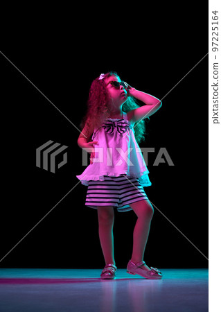 Fashion. Stylish little girl, beginner fashion model wearing sunglasses posing isolated over dark background in neon light. Children emotions, kids fashion style, ad 97225164