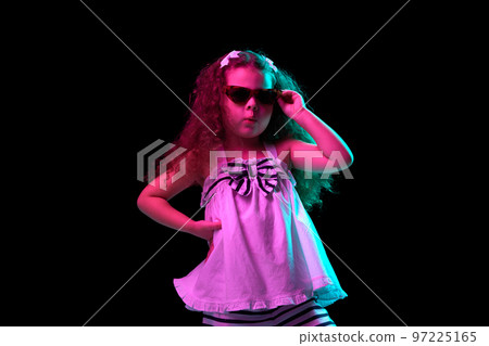 Summer vibes. Stylish little girl, beautiful kid wearing sunglasses posing isolated over dark background in neon light. Children emotions, kids fashion style, ad 97225165