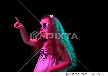 Stylish little girl, beginner fashion model wearing sunglasses posing isolated over dark background in neon light. Children emotions, kids fashion style, ad 97225166