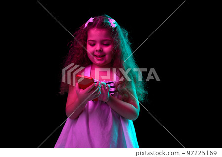 Stylish little girl, beginner fashion model using phone isolated over dark background in neon light. Children emotions, kids fashion style, ad 97225169