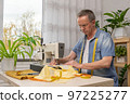 Senior man seamstress sitting and sews on sewing machine in studio. 97225277