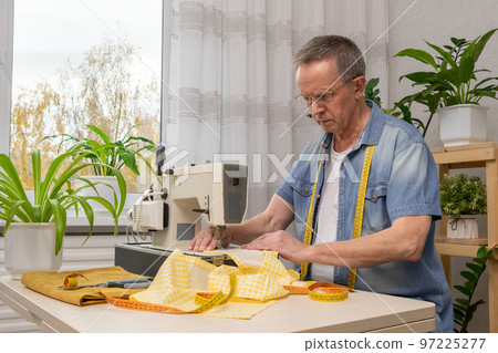 Senior man seamstress sitting and sews on sewing machine in studio. Senior man seamstress sitting and sews on sewing machine in studio. 97225277