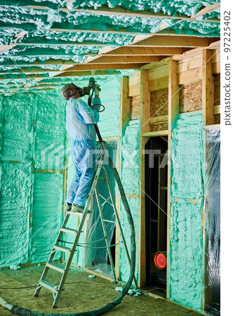 Male builder insulating wooden frame house. Man worker spraying polyurethane foam inside of future cottage, standing on ladder, using plural component gun. Construction and insulation concept. Male builder insulating wooden frame house. Man worker spraying polyurethane foam inside of future cottage, standing on ladder, using plural component gun. Construction and insulation concept. 97225402