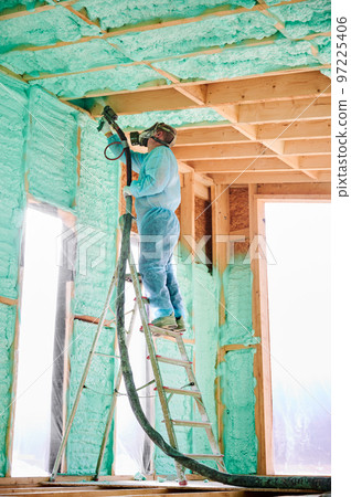 Male builder insulating wooden frame house. Man worker spraying polyurethane foam inside of future cottage, standing on ladder, using plural component gun. Construction and insulation concept. 97225406