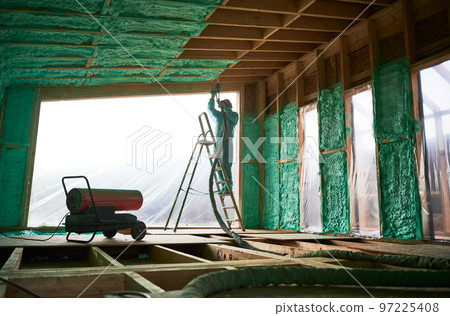 Male builder insulating wooden frame house. Man worker spraying polyurethane foam inside of future cottage, standing on ladder, using plural component gun. Construction and insulation concept. 97225408