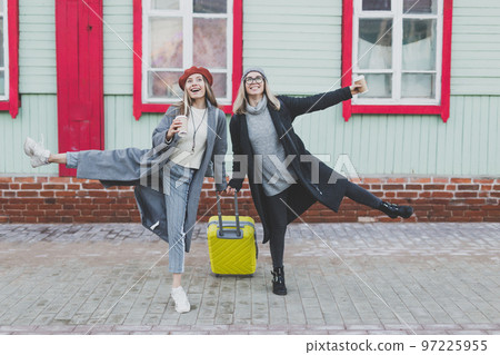Two cheerful funny tourist women smiling and fool around and with suitcases on city street in autumn or spring time - travel and vacation concept Two cheerful funny tourist women smiling and fool around and with suitcases on city street in autumn or spring time - travel and vacation concept 97225955