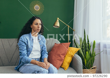 Portrait of sad woman alone at home, pensive Hispanic woman sitting on sofa in living room. 97226290