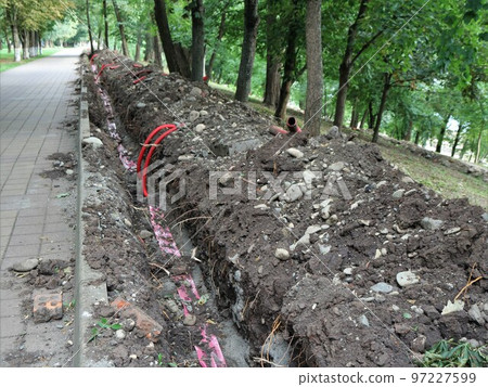 works in a city park on laying electrical communications underground, a trench along a paved path in a park with red plastic pipes with wires for installing lighting 97227599