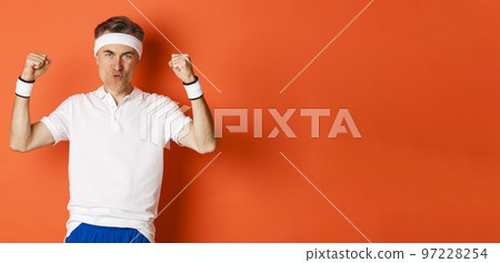 Image of confident middle-aged male athlete, feeling good while working out, raising hands and triumphing, standing over orange background Image of confident middle-aged male athlete, feeling good while working out, raising hands and triumphing, standing over orange background 97228254